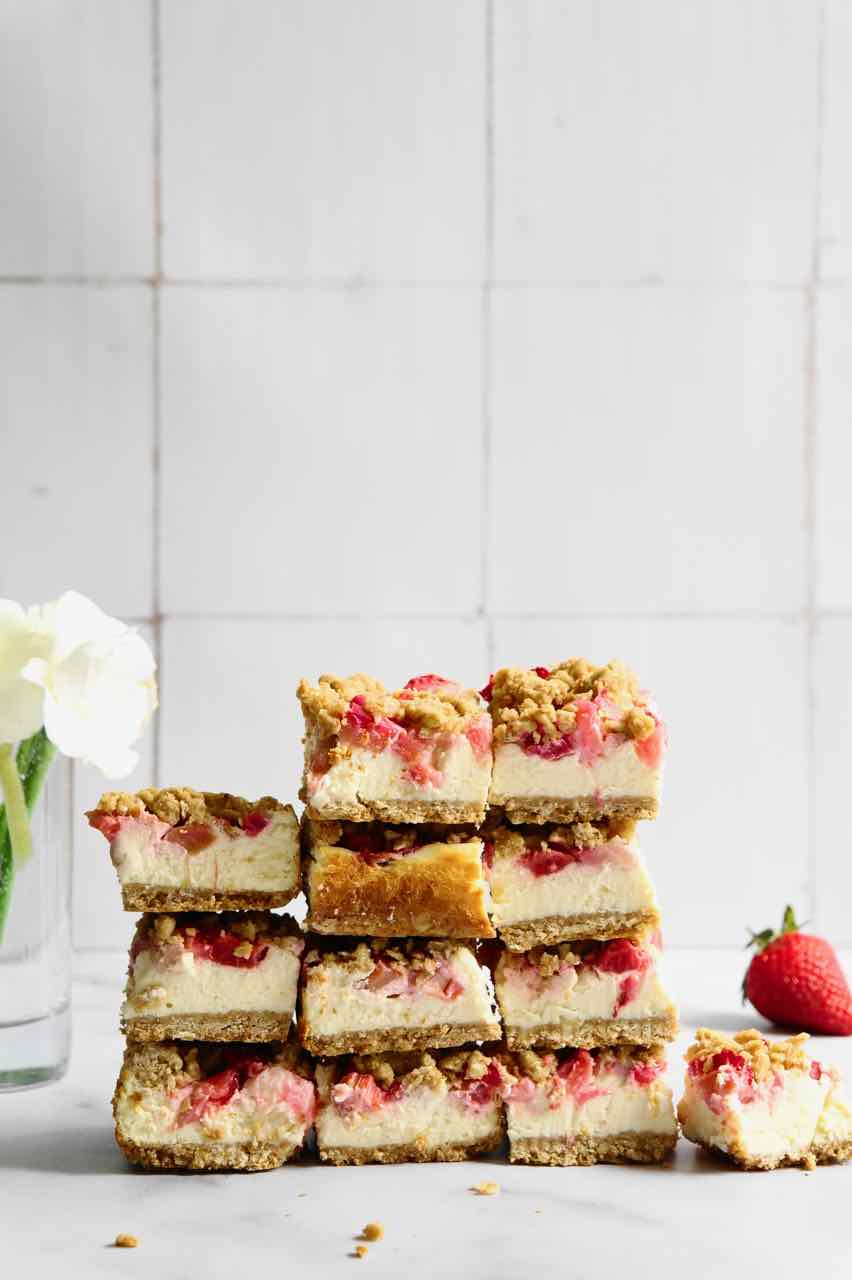 Cheesecake bars with red fruit and streusel piled in stacks in front of a white tile backsplash.